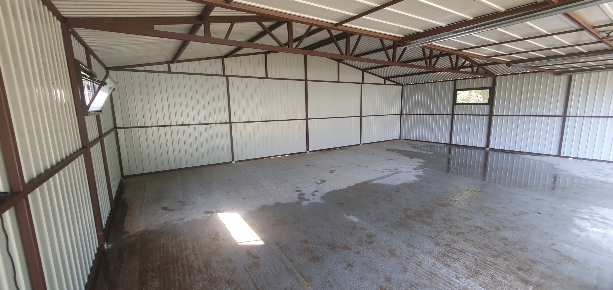 Wnętrze nowego, pustego garażu blaszanego z oknem, z wilgotną podłogą betonową odbijającą światło słoneczne.