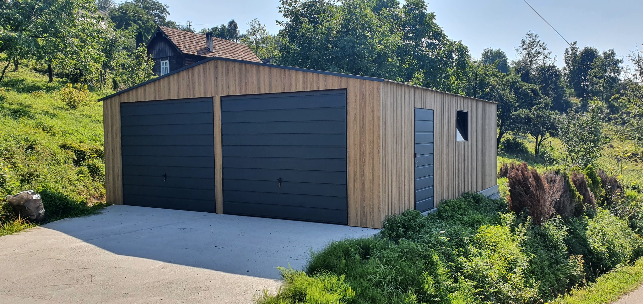 Nowoczesny, dwustanowiskowy garaż z elewacją imitującą drewno i antracytowymi bramami segmentowymi, otoczony zielenią i z widokiem na dom w tle.
