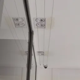 Biały sufit z zamontowanymi dwoma podwójnymi reflektorami halogenowymi, pojedynczym reflektorem oraz kamerą przemysłową, widoczne przewody zasilające, odbicie w szklanej powierzchni.
