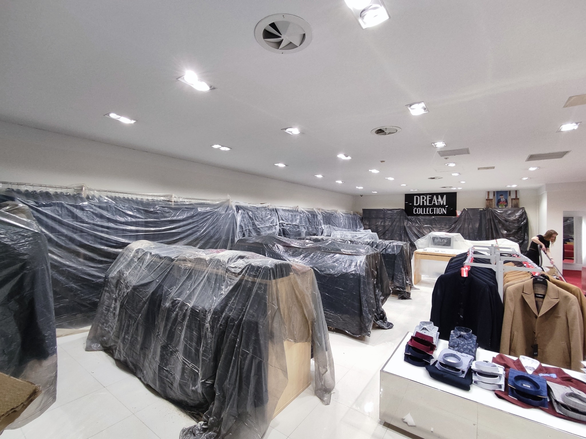 Sklep odzieżowy w trakcie remontu lub zabezpieczania towaru przed remontem: meble i ubrania okryte folią ochronną, widoczne oświetlenie sufitowe i wentylacja.