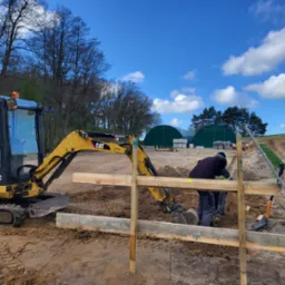 Żółta mini koparka CAT 301.8C pracuje przy wykopie fundamentów, widoczni dwaj pracownicy w szarych spodniach i ciemnych kurtkach, drewniane szalunki, w tle zielone hale i błękitne niebo z białymi...