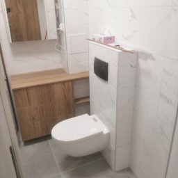 Michal Zientarski - Nowoczesna toaleta podwieszana z zabudową, białe płytki imitujące marmur, drewniana szafka pod umywalką z szufladami, szara podłoga, pudełko chusteczek na obudowie stelaża.