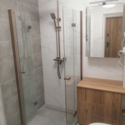 Michal Zientarski - Nowoczesna łazienka z kabiną prysznicową typu walk-in, armaturą w kolorze miedzianym i szafką pod umywalkę z drewna o widocznej strukturze słojów.