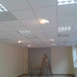 Michal Zientarski - Montaż sufitu podwieszanego kasetonowego w biurze, widoczny pracownik przy ścianie, drabina i częściowo opuszczona roleta.