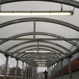 Michal Zientarski - Zadaszony chodnik z metalową konstrukcją łukową i oświetleniem jarzeniowym, widoczna osoba w oddali.