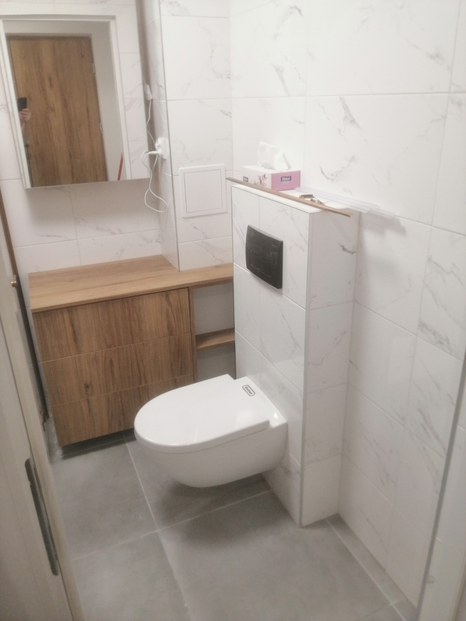 Nowoczesna toaleta podwieszana z zabudową, białe płytki imitujące marmur, drewniana szafka pod umywalką z szufladami, szara podłoga, pudełko chusteczek na obudowie stelaża.