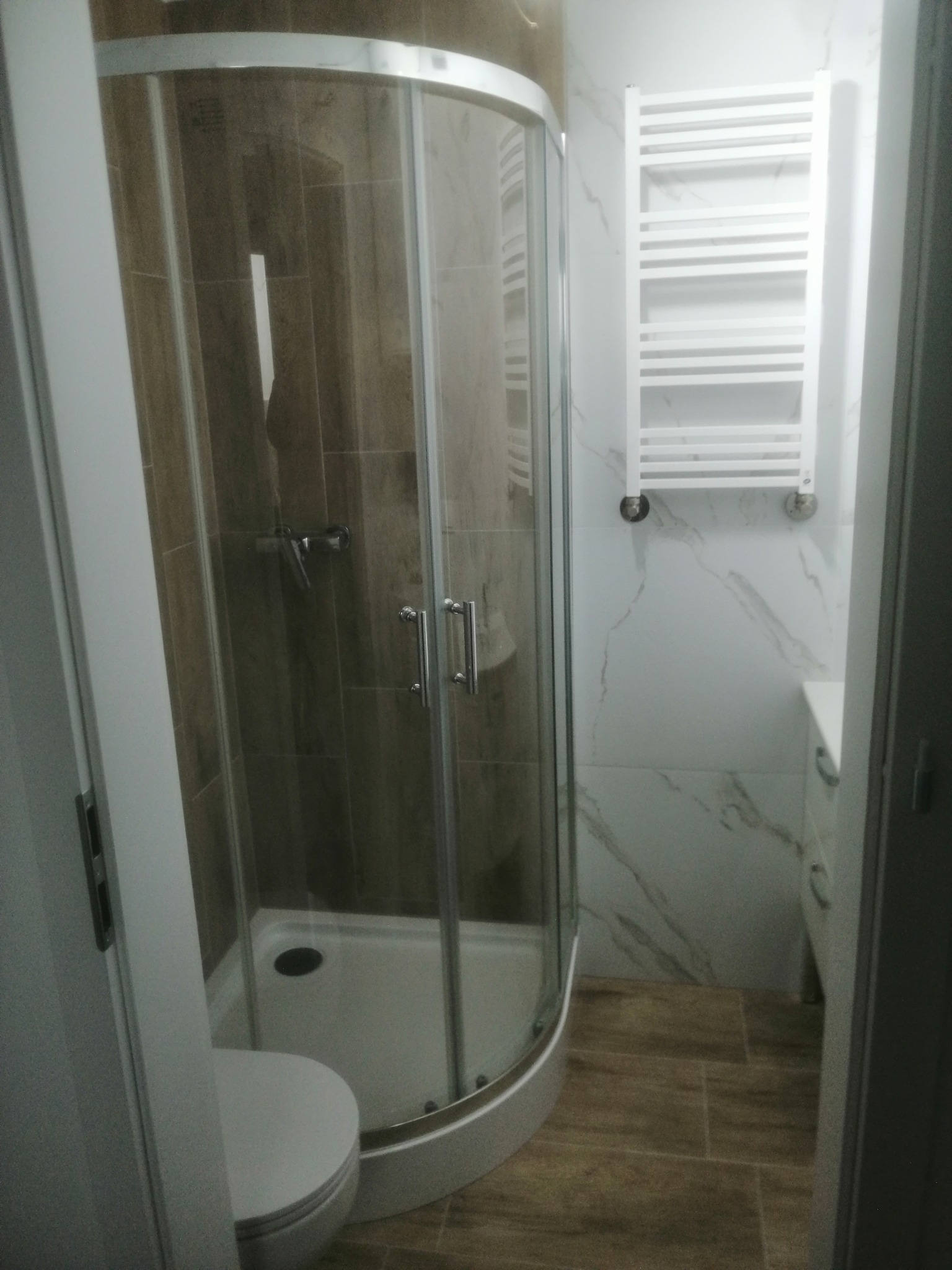 Nowoczesna kabina prysznicowa narożna z zaokrąglonymi drzwiami w łazience wykończonej płytkami imitującymi drewno i marmur, widoczny biały grzejnik drabinkowy.