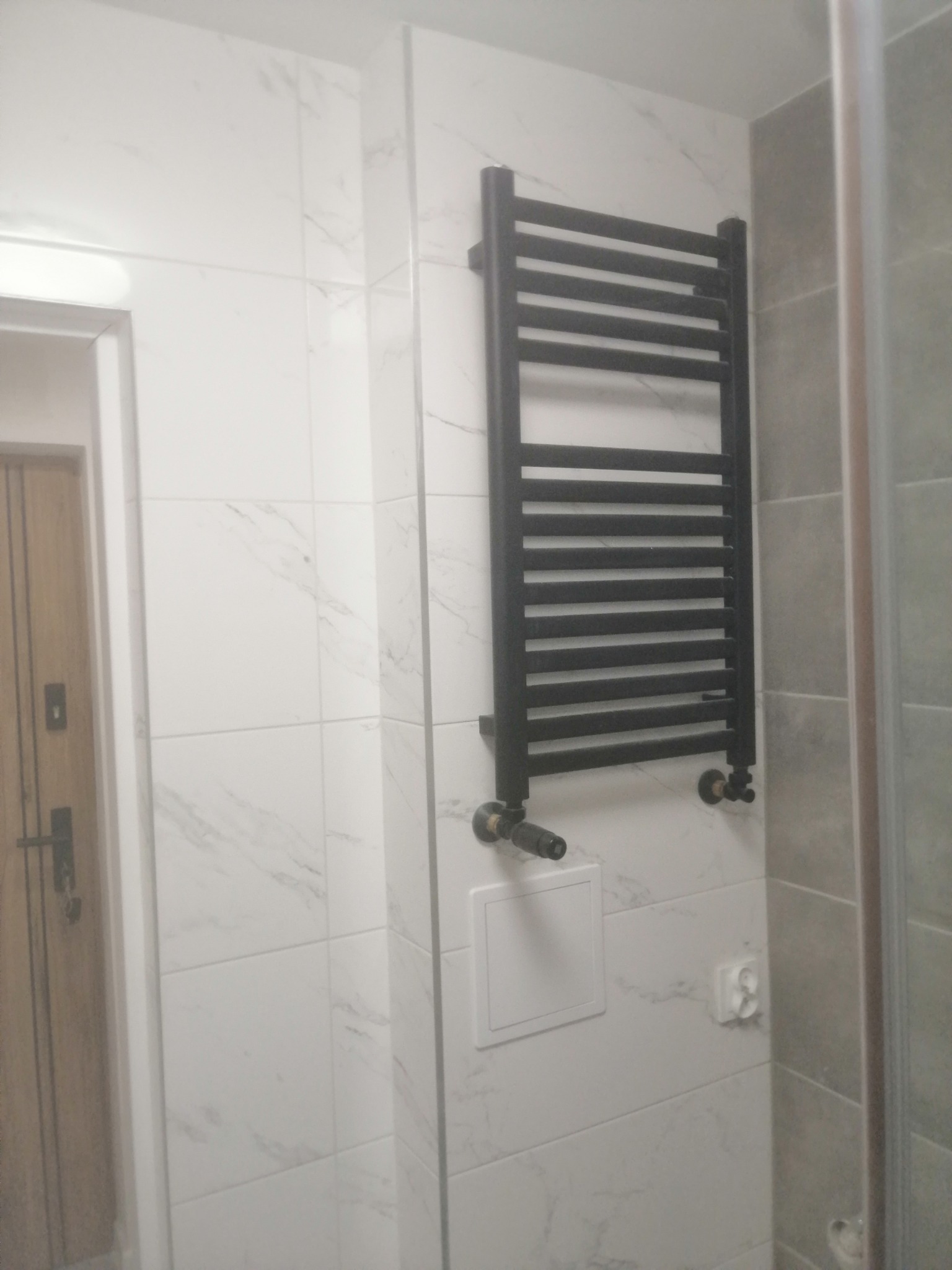 Łazienka po remoncie z grzejnikiem drabinkowym w kolorze czarnym na ścianie z białych płytek imitujących marmur, widoczny fragment kabiny prysznicowej i drzwi.