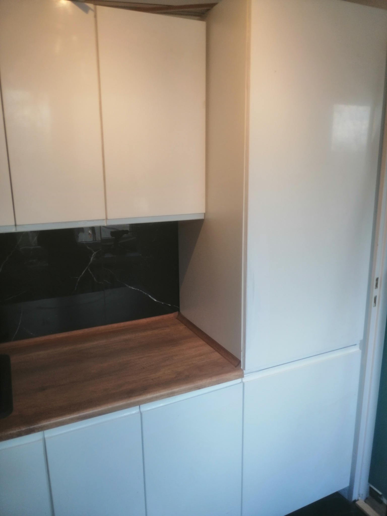 Białe, lakierowane szafki kuchenne z drewnianym blatem i czarnym panelem nad blatem imitującym marmur.