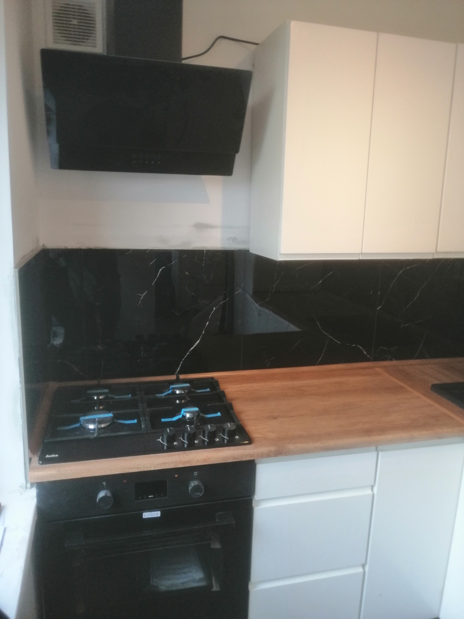 Nowoczesna kuchnia z czarnym panelem ściennym imitującym marmur, białymi szafkami i drewnianym blatem, widoczny czarny okap i płyta gazowa z piekarnikiem.