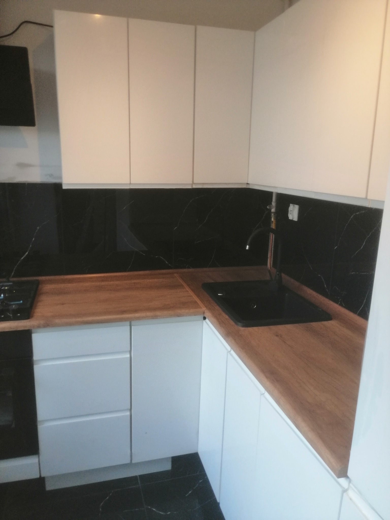 Nowoczesna kuchnia z białymi szafkami, drewnianym blatem i czarnym marmurowym backsplashiem. Czarny zlew i bateria. Widoczna płyta grzewcza po lewej.