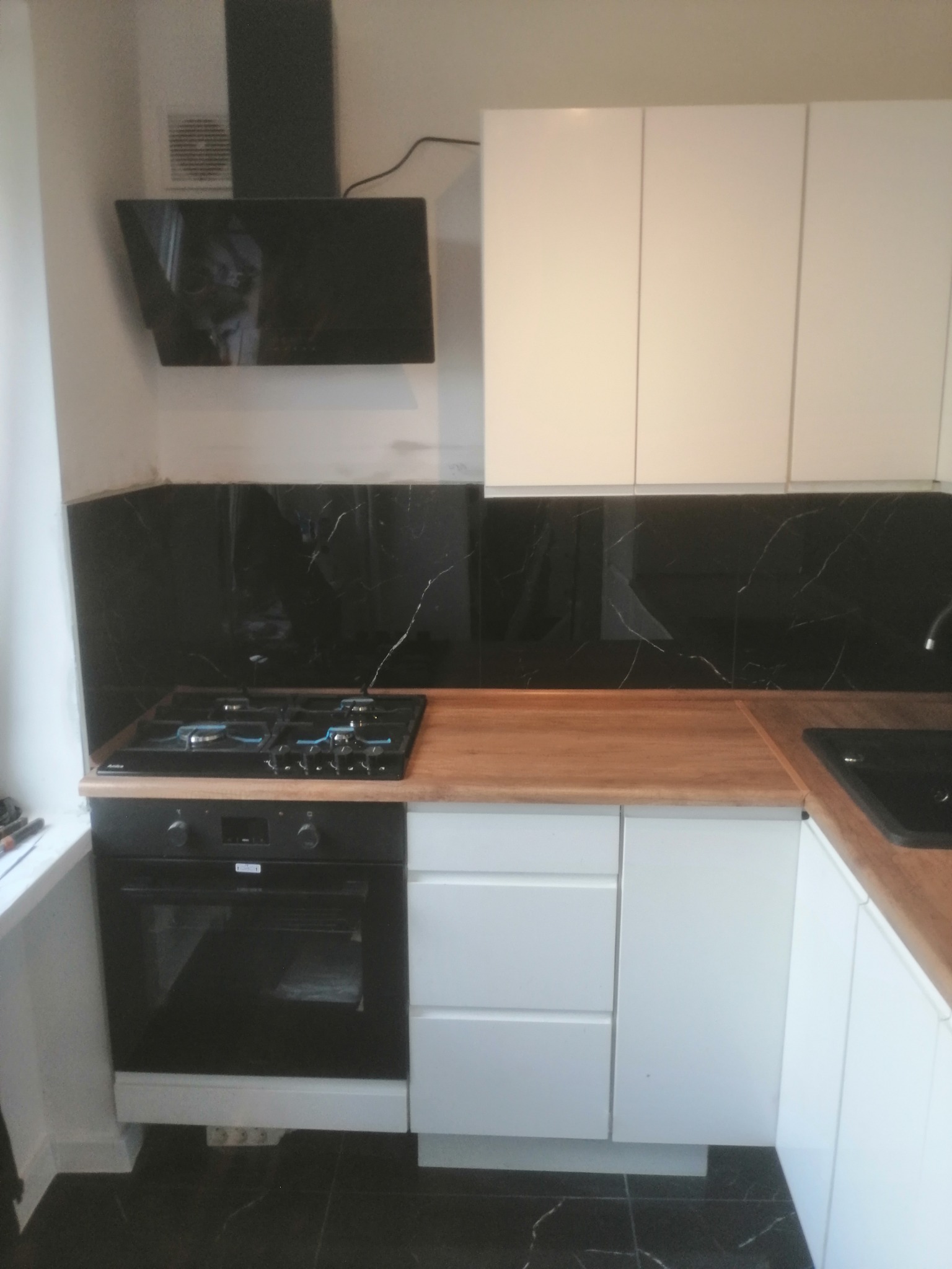 Wykończona kuchnia z białymi szafkami, drewnianym blatem, czarnym panelem nad blatem imitującym marmur i czarną podłogą.