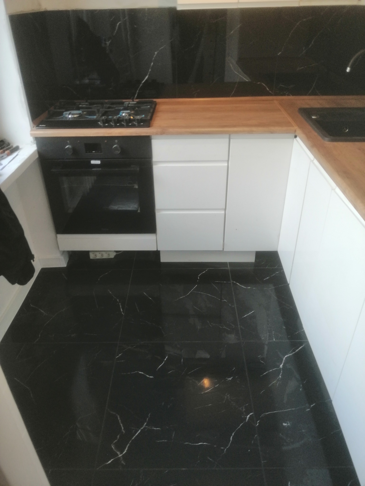 Częściowy widok kuchni po remoncie: czarne płytki podłogowe i na ścianie nad blatem, białe szafki, drewniany blat, czarny piekarnik i płyta gazowa.