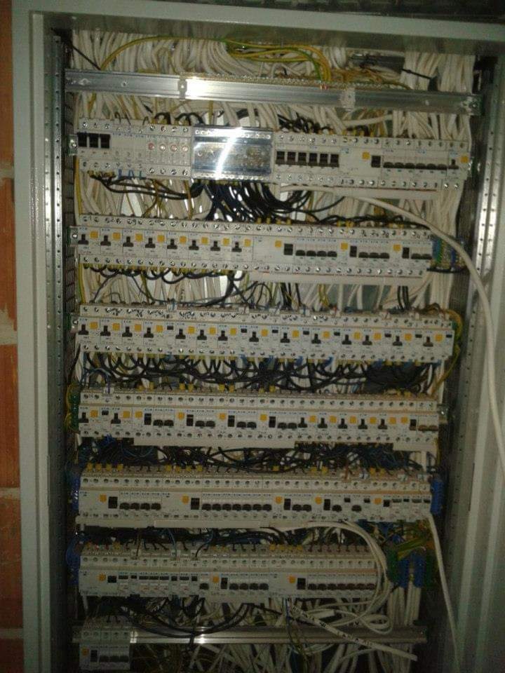 Rozdzielnica elektryczna z wieloma zabezpieczeniami, widoczne okablowanie w metalowej szafie, ceglana ściana obok.