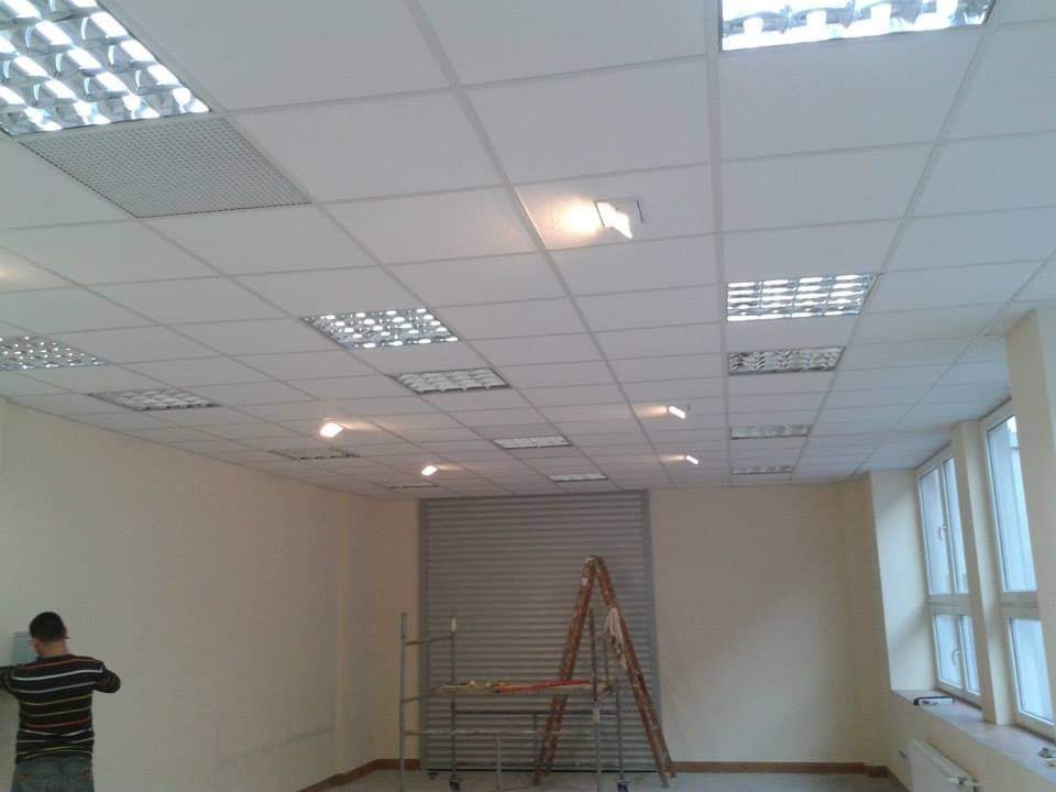 Montaż sufitu podwieszanego kasetonowego w biurze, widoczny pracownik przy ścianie, drabina i częściowo opuszczona roleta.