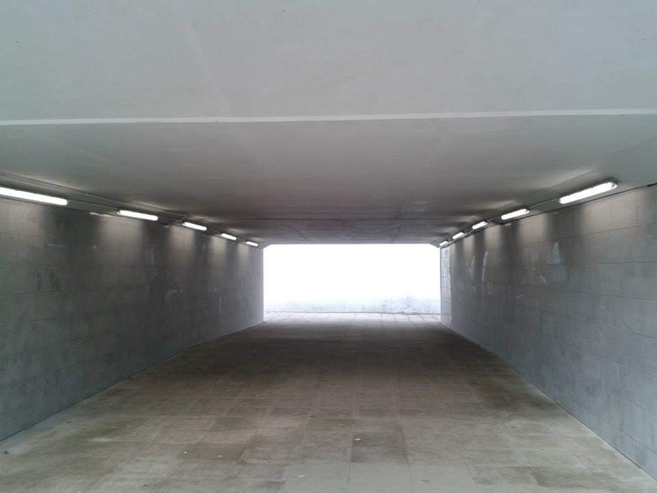 Betonowy tunel dla pieszych z rzędami lamp fluorescencyjnych po obu stronach, prowadzący do jasnego wyjścia.