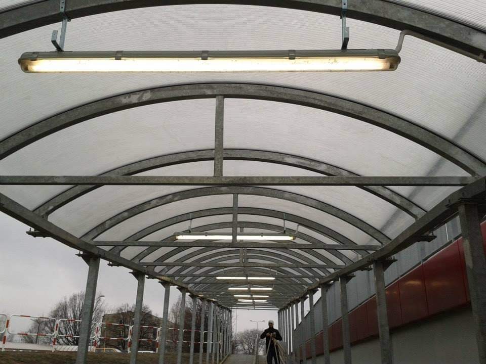 Zadaszony chodnik z metalową konstrukcją łukową i oświetleniem jarzeniowym, widoczna osoba w oddali.