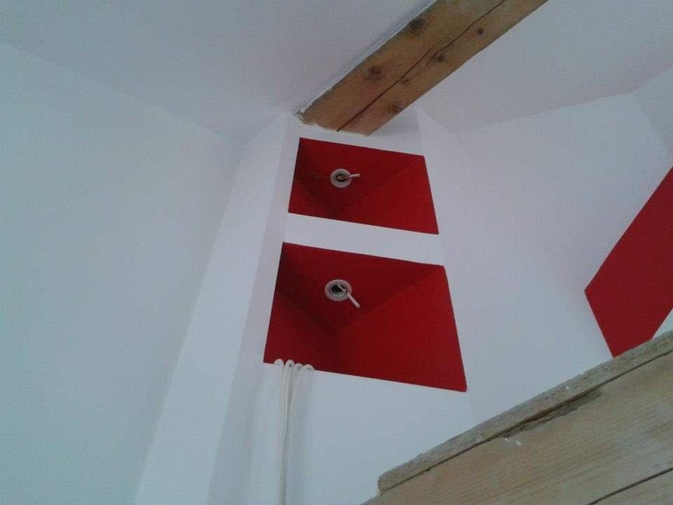 Biała ściana z dwoma czerwonymi, trapezoidalnymi panelami z metalowymi oczkami i wystającymi przewodami, widoczne fragmenty drewnianych belek stropowych.