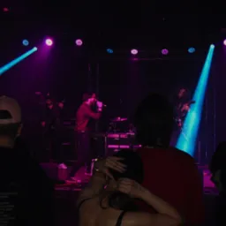 Zespół na scenie podczas koncertu w klubie, oświetlony niebieskimi i fioletowymi reflektorami, widoczni muzycy grający na gitarach i śpiewający do mikrofonu, w tle perkusja, na pierwszym planie...