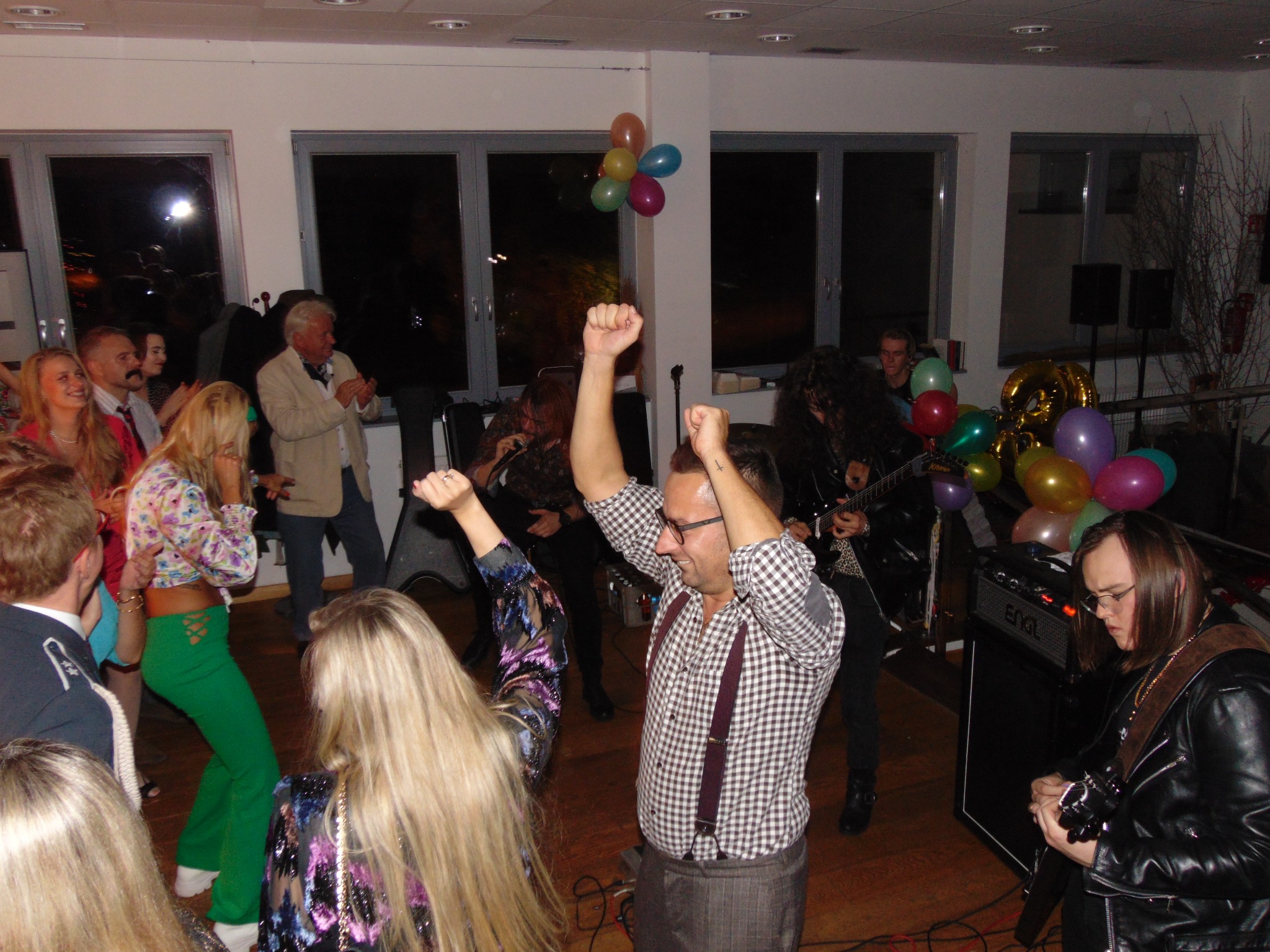 Zespół grający na żywo podczas imprezy urodzinowej: wokalista w okularach i szelkach, gitarzyści w skórzanych kurtkach, tańczący goście, balony w tle.