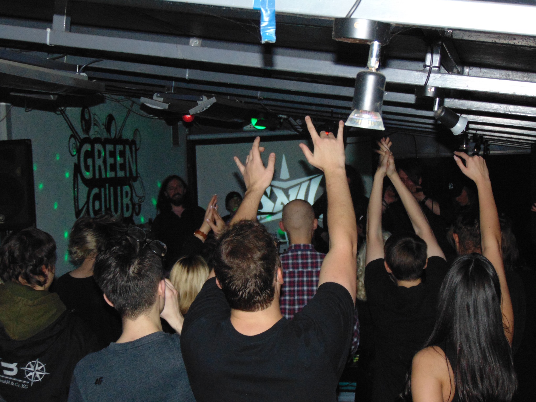 Publiczność z podniesionymi rękami podczas koncertu w klubie Green Club, widoczny fragment sceny z logo klubu i oświetleniem.