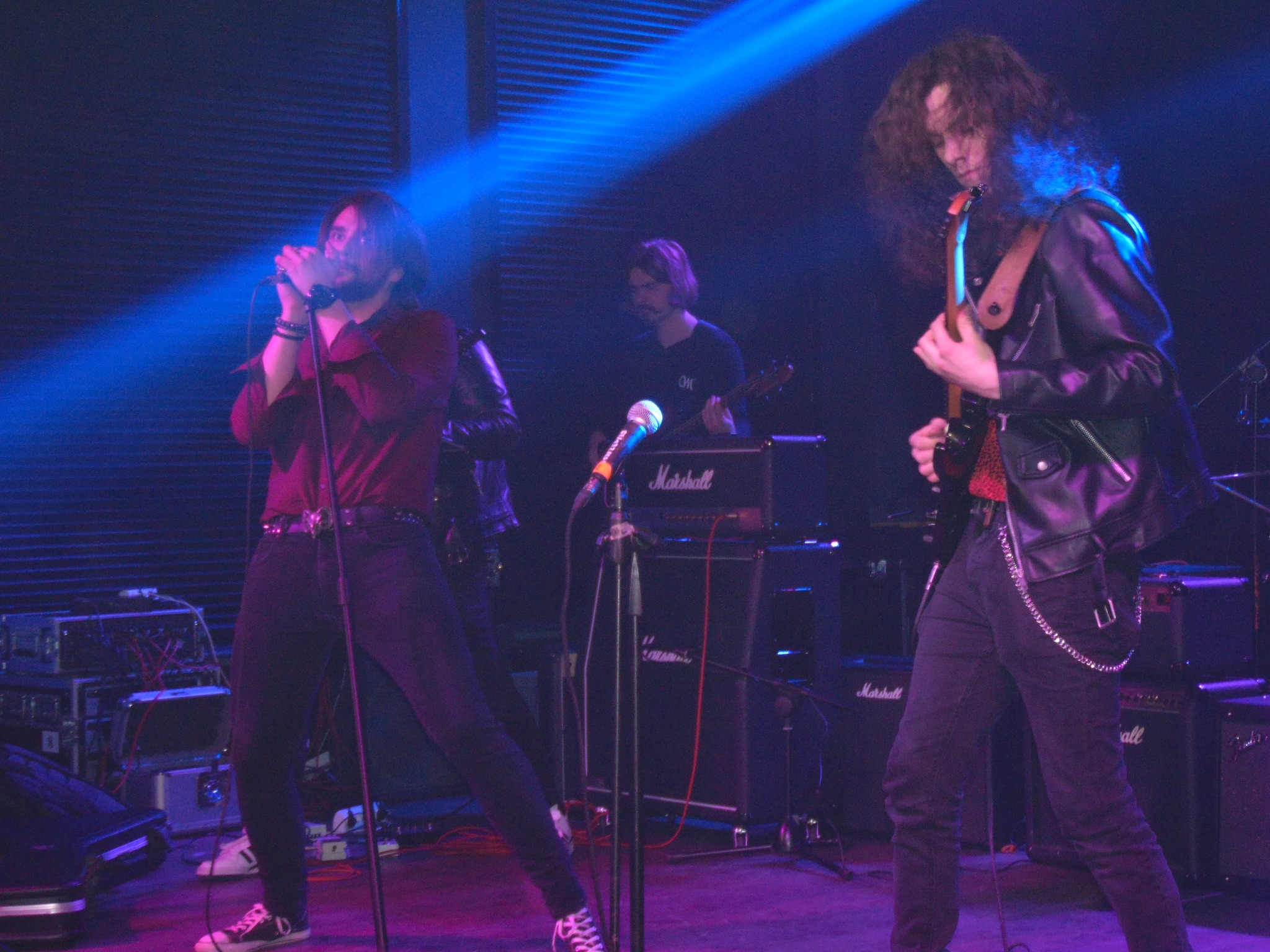 Wokalista w bordowej koszuli śpiewa do mikrofonu na scenie oświetlonej niebieskim światłem, obok gitarzysta w skórzanej kurtce gra na gitarze elektrycznej, w tle widoczny basista i wzmacniacze...