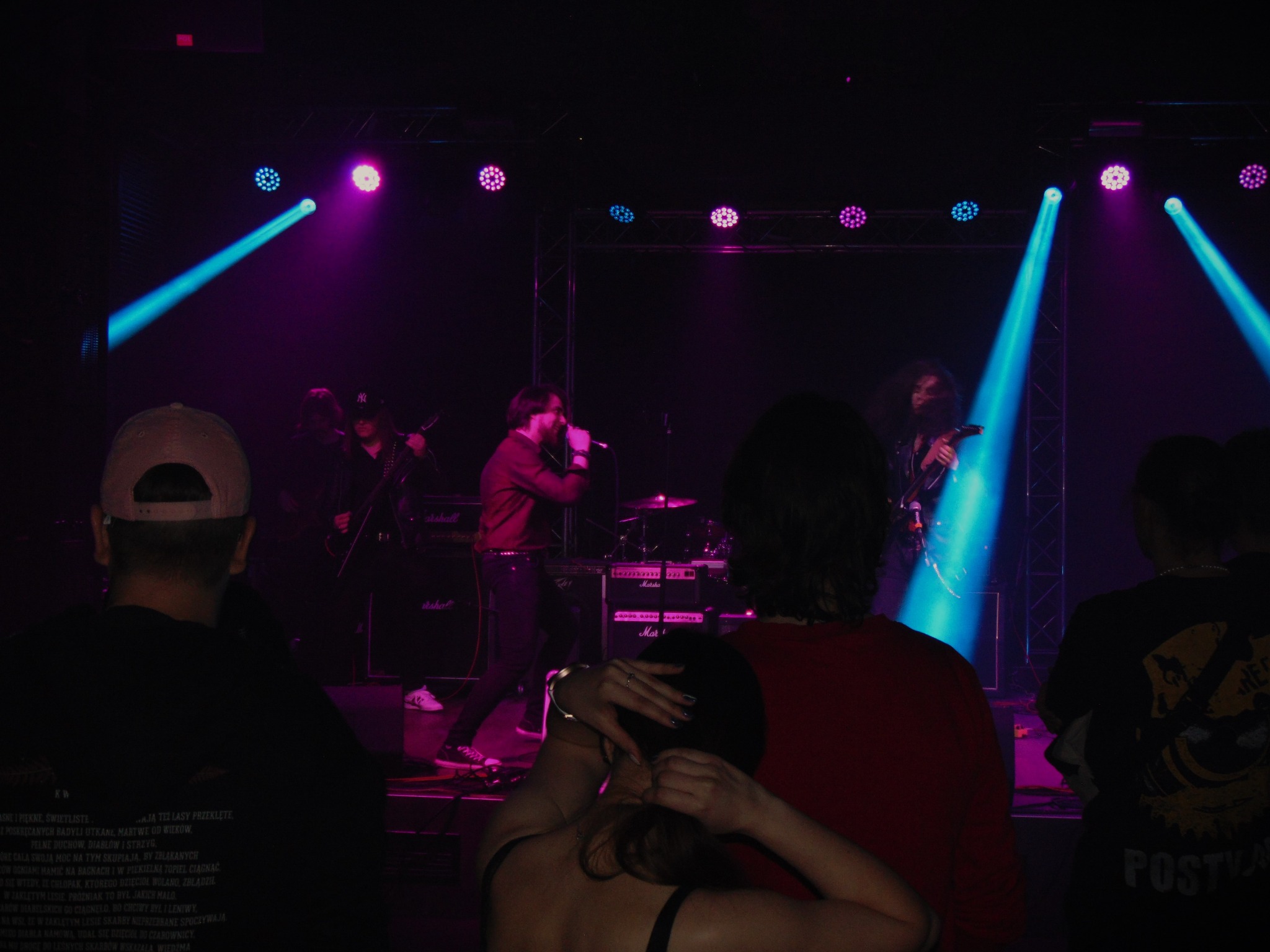 Zespół na scenie podczas koncertu w klubie, oświetlony niebieskimi i fioletowymi reflektorami, widoczni muzycy grający na gitarach i śpiewający do mikrofonu, w tle perkusja, na pierwszym planie...