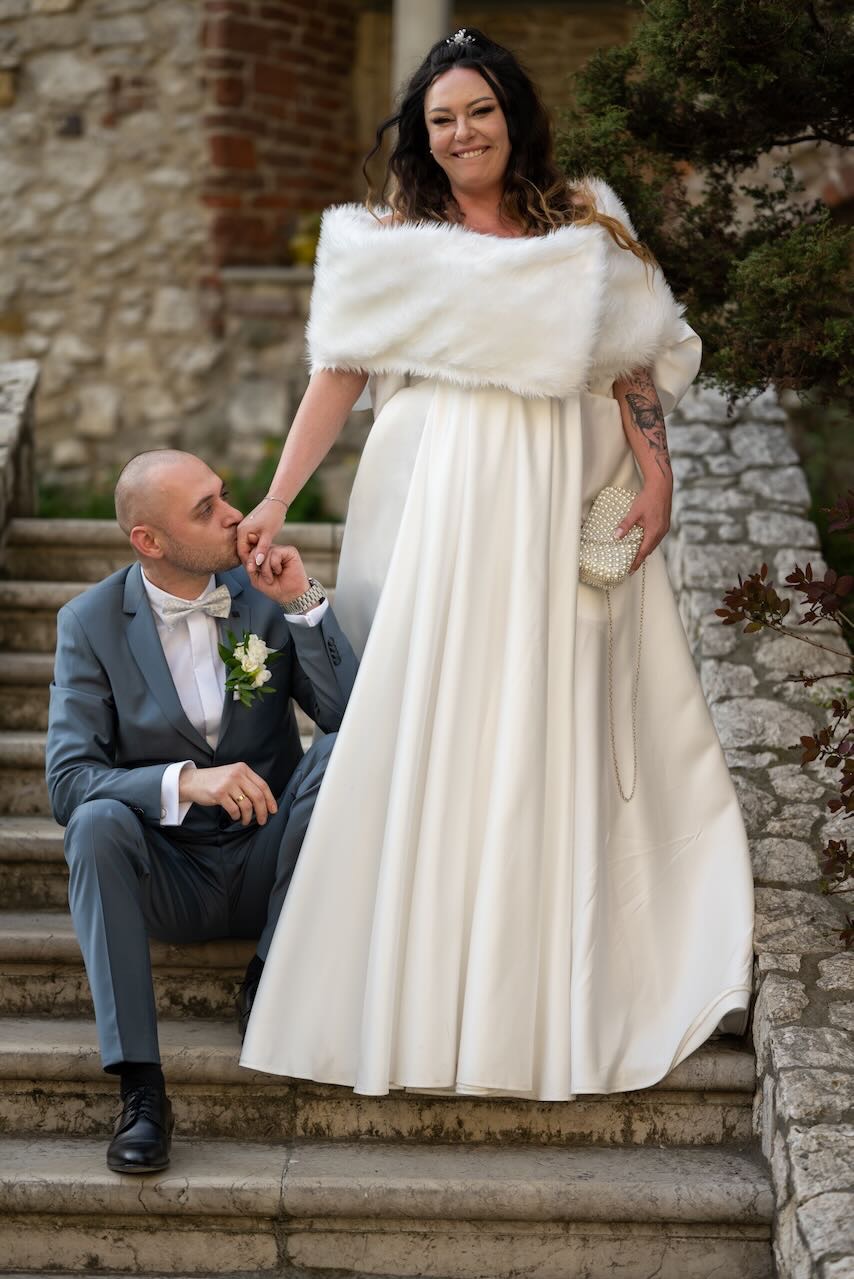 Pan młody w szarym garniturze całuje dłoń panny młodej w białej sukni z futrzanym szalem i torebką z pereł na kamiennych schodach.