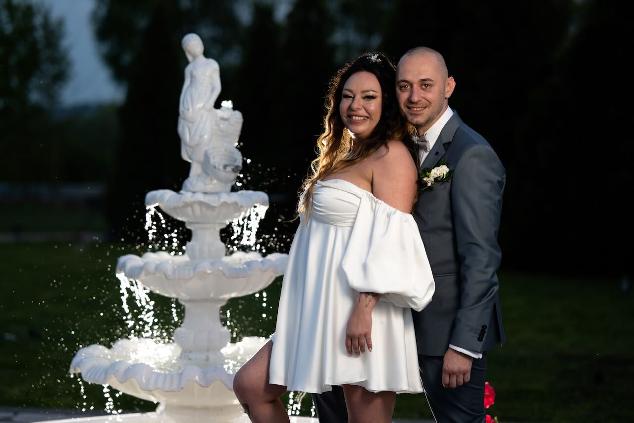 Para młoda uśmiechnięta, pozująca obok wielopoziomowej, oświetlonej fontanny z rzeźbą, panna młoda w krótkiej białej sukience z odkrytymi ramionami, pan młody w szarym garniturze z butonierką.