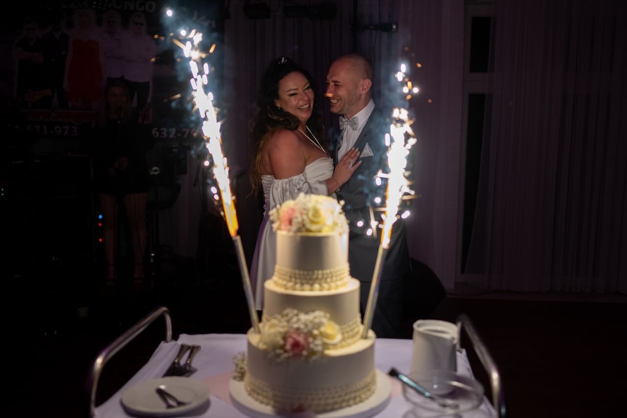 Uśmiechnięta para młoda za trzypiętrowym tortem weselnym z kwiatami, otoczona fontannami iskier.