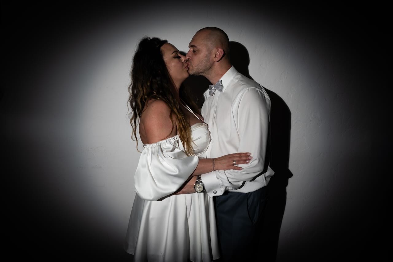 Para młoda całująca się na tle ściany z efektem światłocienia, panna młoda w krótkiej białej sukience z odkrytymi ramionami, pan młody w białej koszuli z muchą i zegarkiem na ręku.