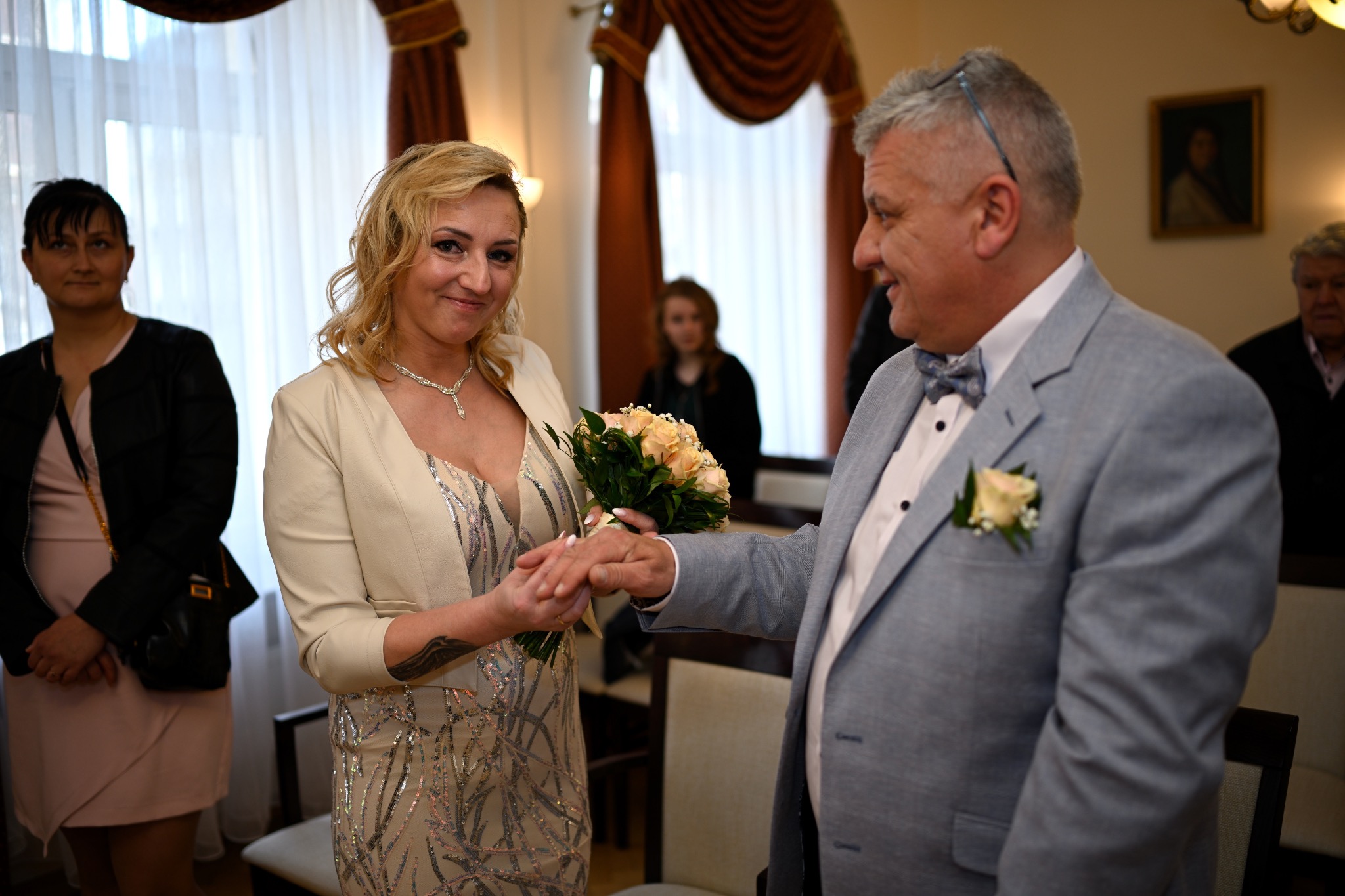 Nowożeńcy wymieniają się obrączkami podczas ceremonii ślubnej w sali z portretem na ścianie i osobami w tle.