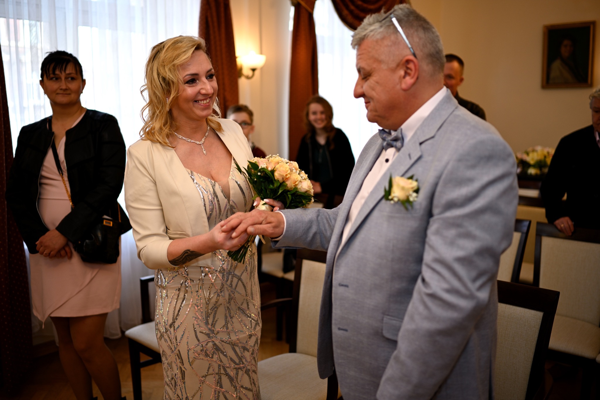 Panna młoda w sukni z cekinami i kremowym żakiecie trzyma bukiet róż, a pan młody w szarym garniturze wkłada jej obrączkę podczas ceremonii ślubnej w pomieszczeniu z gośćmi w tle.
