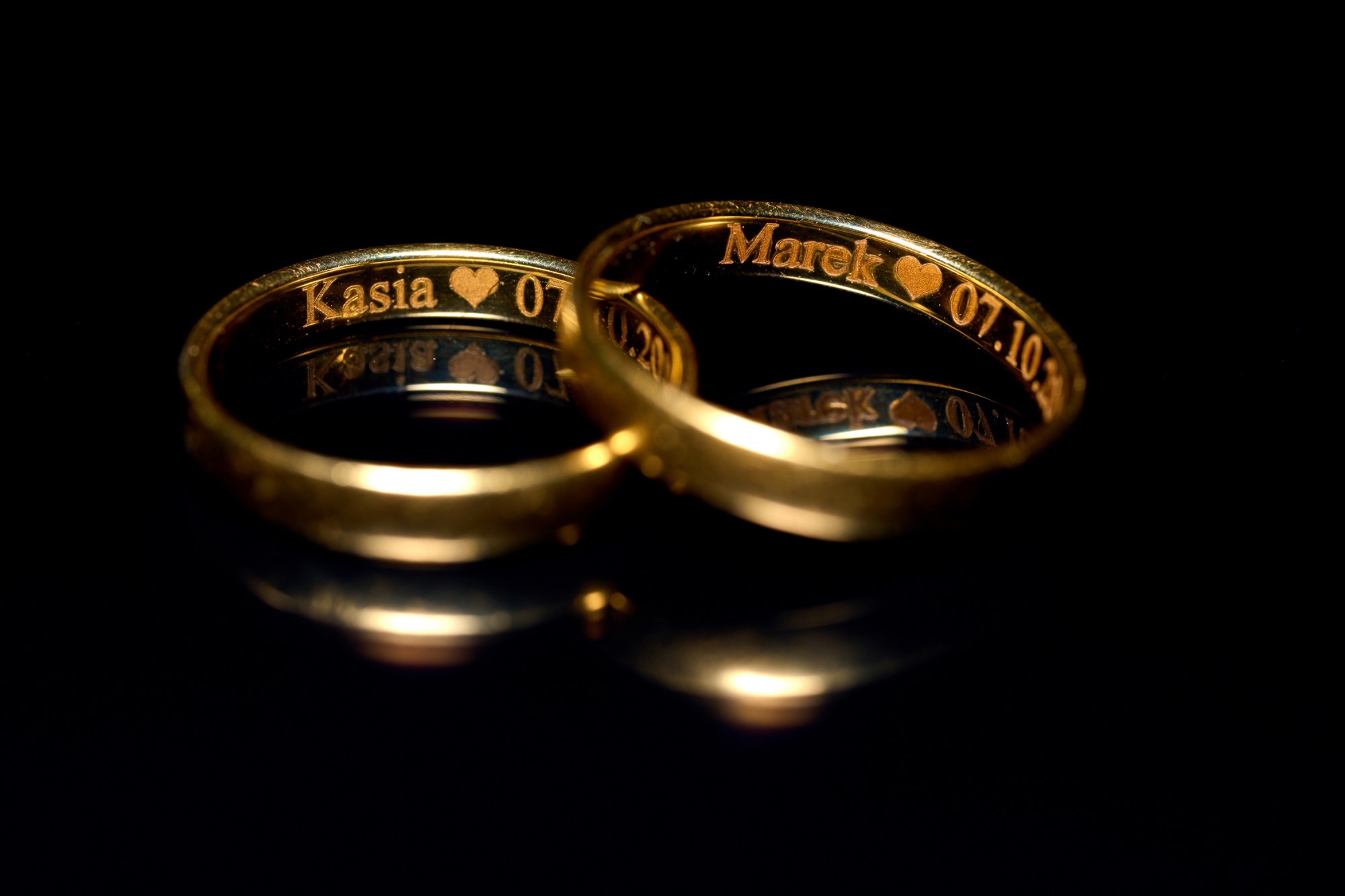 Dwa złote obrączki ślubne z wygrawerowanymi imionami Kasia i Marek oraz datą 07.10.20, leżące na czarnym, błyszczącym tle, tworzącym lustrzane odbicie.