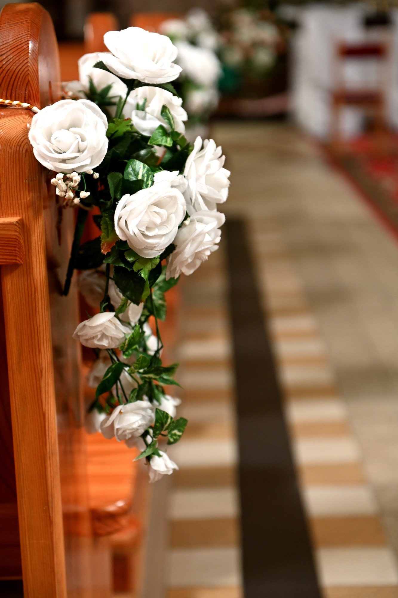 Dekoracja kościelnej ławki z białych róż i zielonych liści, widok na korytarz z czerwonym dywanem w tle.