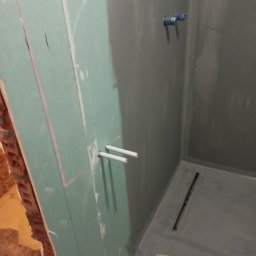 Janusz Szmyt - Przygotowanie do montażu kabiny prysznicowej: widoczne surowe ściany z płyt kartonowo-gipsowych z zaznaczonymi liniami, wystające rury instalacji wodnej oraz odpływ liniowy w posadzce, całość...