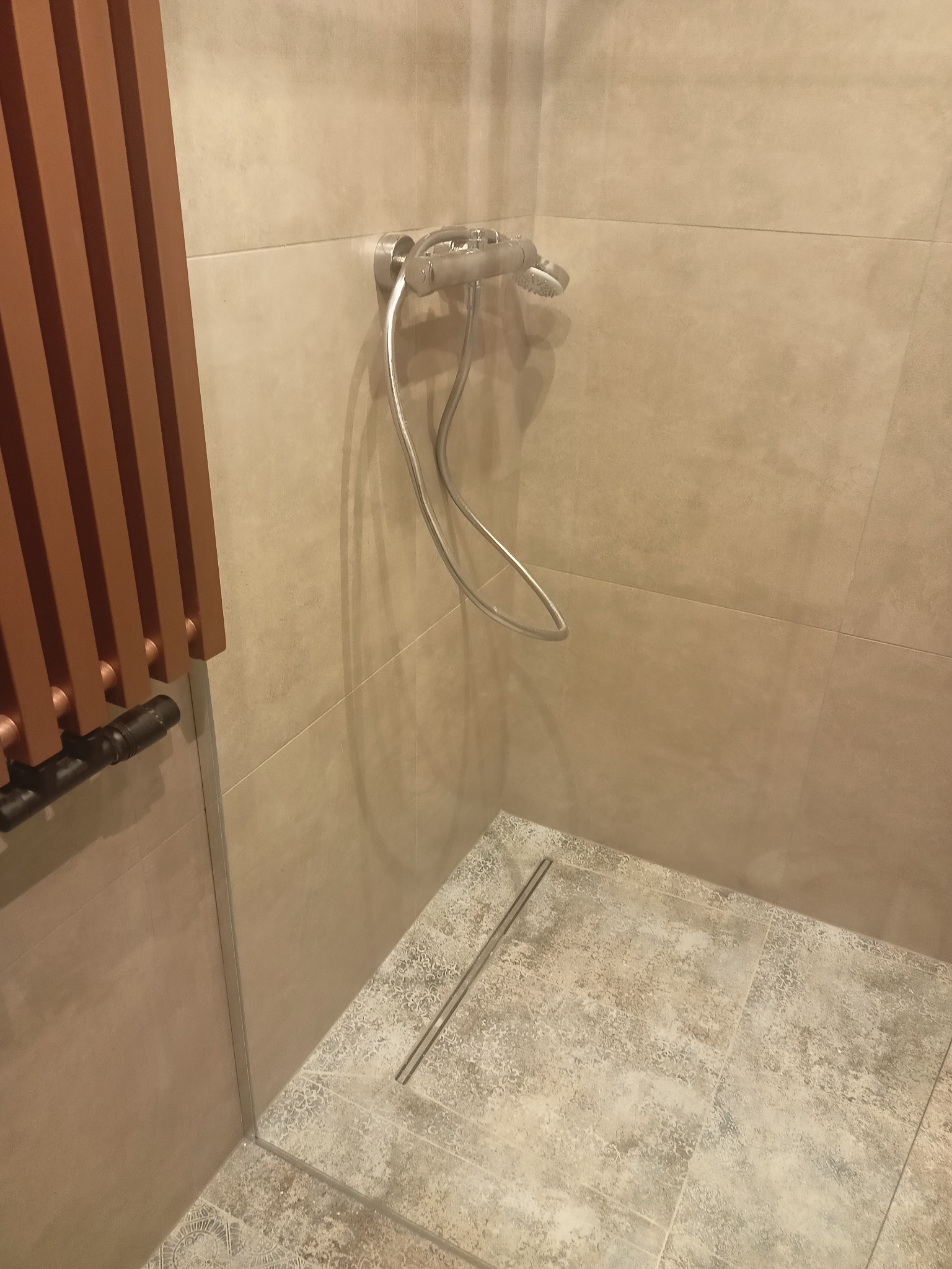 Beżowa łazienka z odpływem liniowym, chromowaną baterią natryskową i grzejnikiem drabinkowym w kolorze miedzianym.