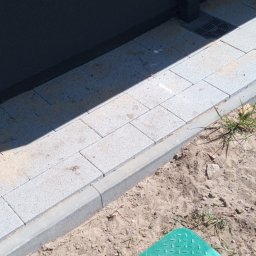 Piotr Oporski - Ułożona szara kostka brukowa przy ścianie budynku, z zieloną pokrywą studzienki rewizyjnej w piasku.