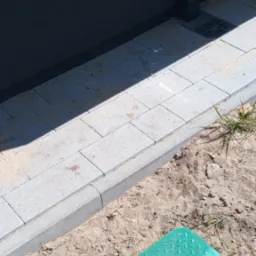 Ułożona szara kostka brukowa przy ścianie budynku, z zieloną pokrywą studzienki rewizyjnej w piasku.