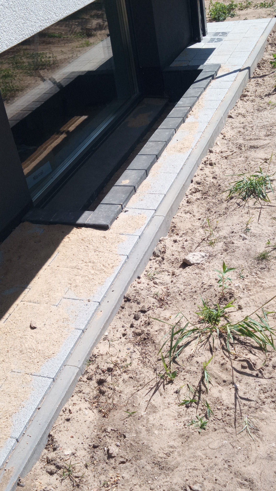 Krawężnik i fragment ułożonej kostki brukowej przy oknie piwnicznym, podsypane piaskiem, widoczne chwasty na ziemi.