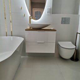 Remonty i wykończenia wnętrz Łukasz Musiał - Nowoczesna łazienka w odcieniach bieli i złota, z owalnym lustrem z podświetleniem LED, wolnostojącą wanną, umywalką nablatową na szafce z drewnianym blatem i wiszącą toaletą.