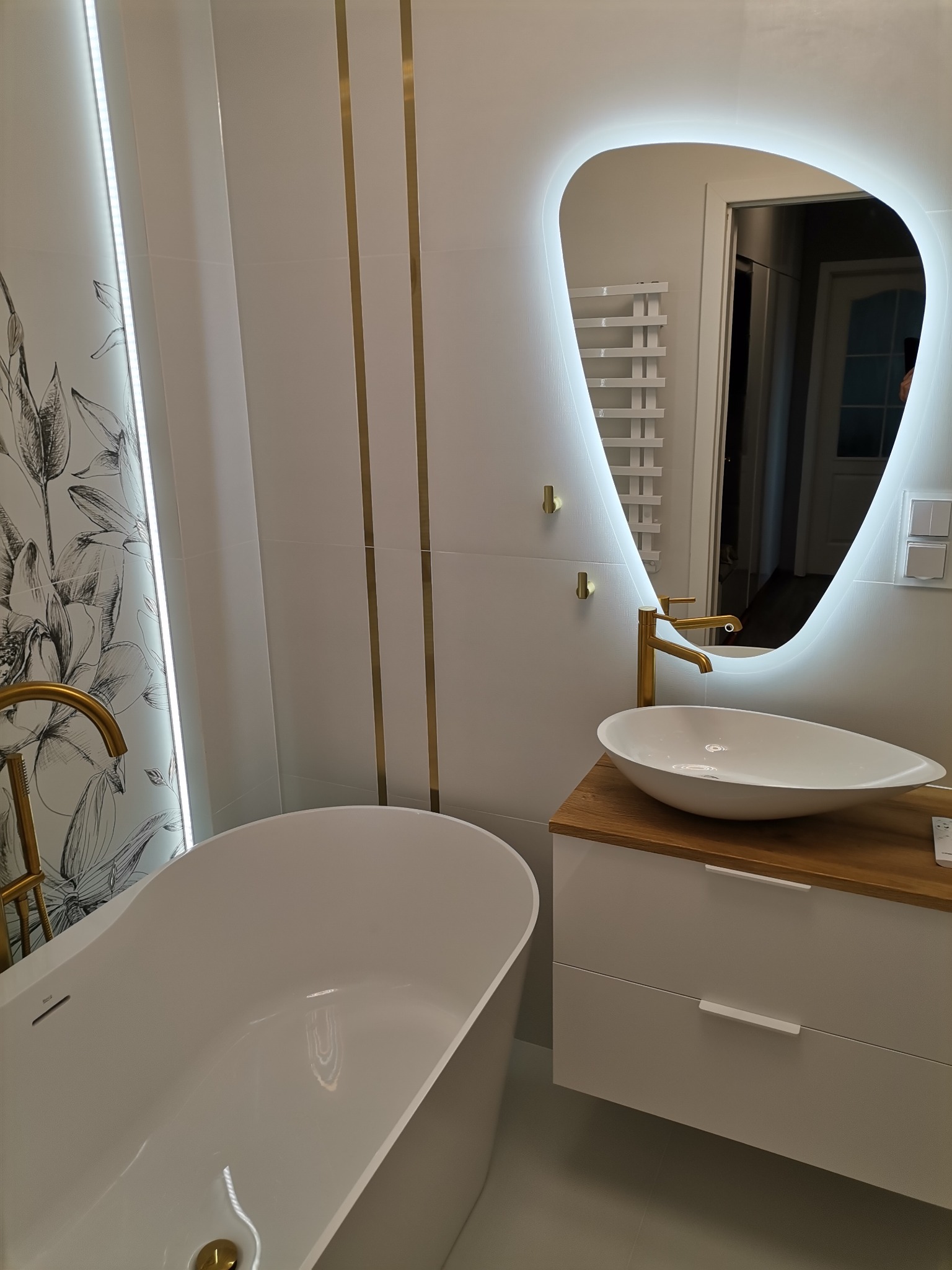 Nowoczesna łazienka z wolnostojącą wanną, umywalką nablatową na szafce z drewnianym blatem i podświetlanym lustrem o nieregularnym kształcie, złote akcenty.