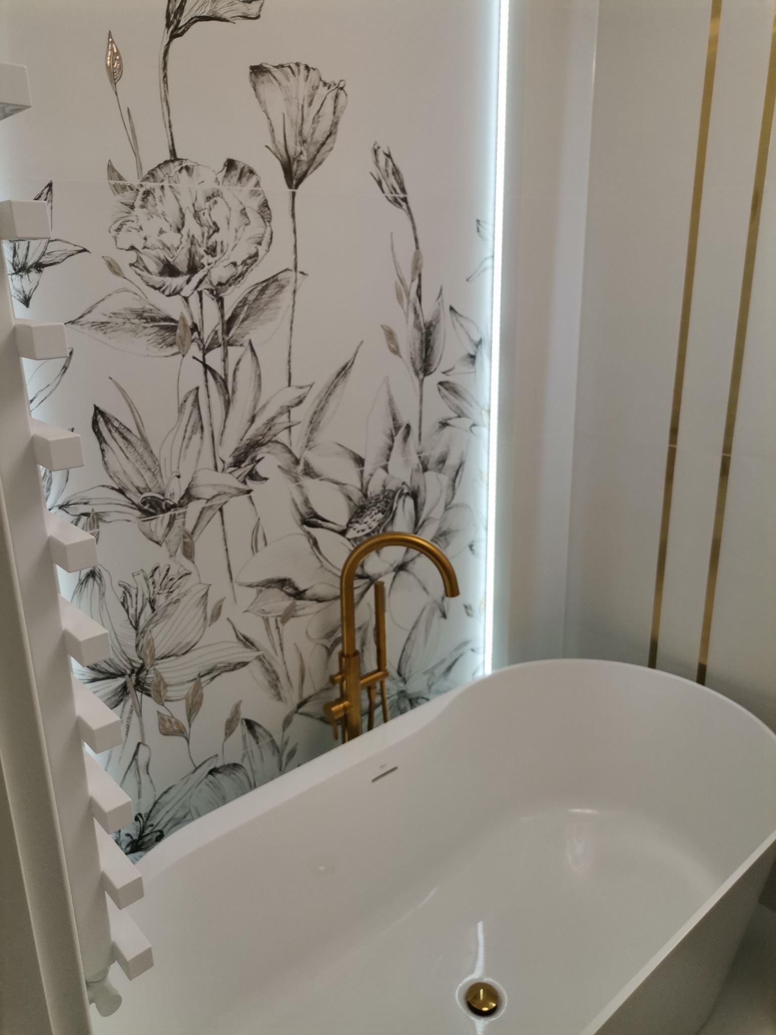 Biała wanna wolnostojąca z złotą armaturą na tle ściany z motywem kwiatowym w odcieniach szarości i bieli, widoczny fragment grzejnika drabinkowego.
