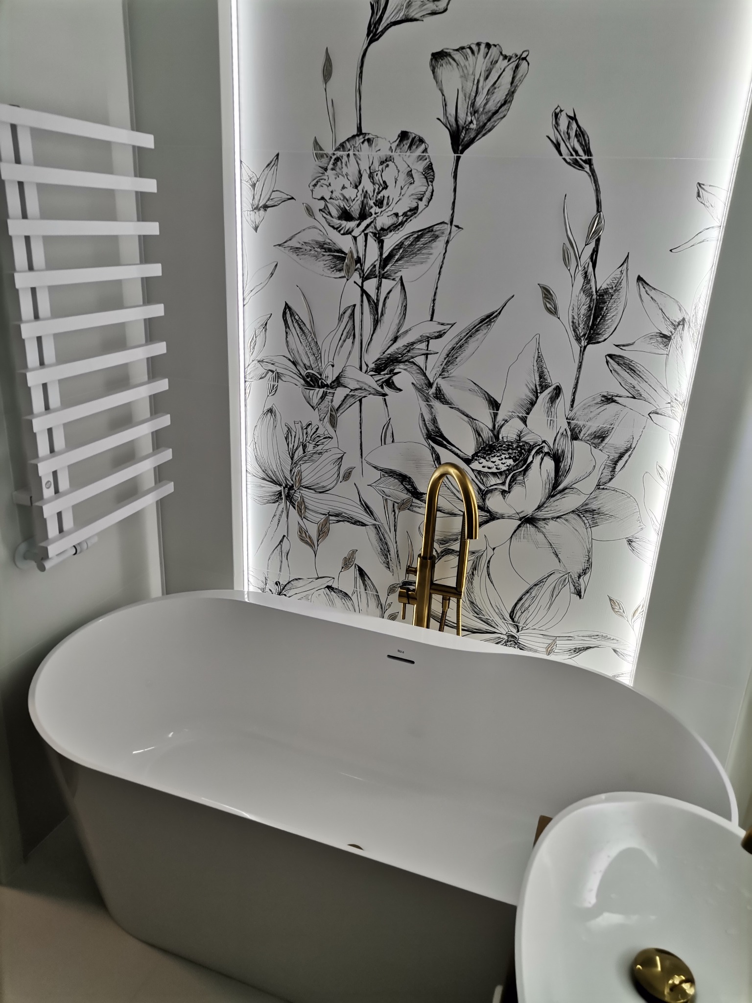 Nowoczesna łazienka z wolnostojącą wanną, złotą armaturą i grzejnikiem drabinkowym na tle ściany z motywem kwiatowym.
