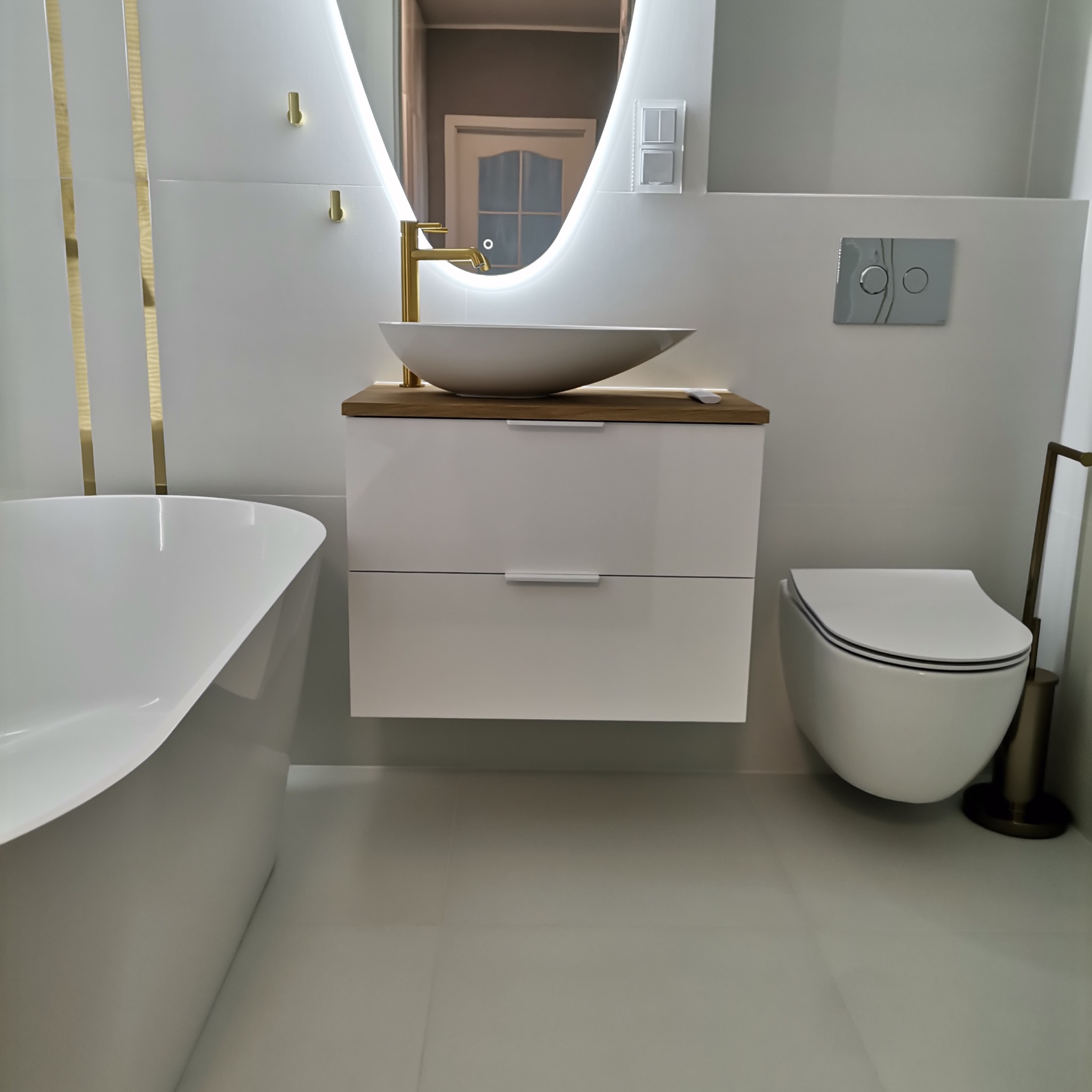 Nowoczesna łazienka w odcieniach bieli i złota, z owalnym lustrem z podświetleniem LED, wolnostojącą wanną, umywalką nablatową na szafce z drewnianym blatem i wiszącą toaletą.