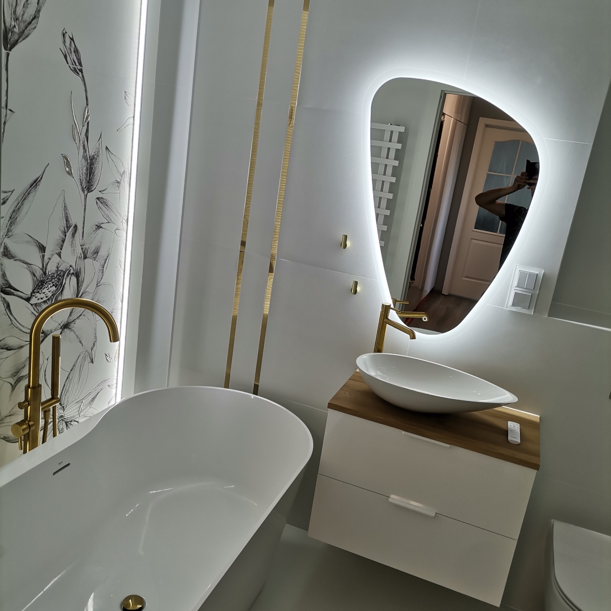 Nowoczesna łazienka z wolnostojącą wanną, złotą armaturą, umywalką nablatową na drewnianym blacie i podświetlanym lustrem o nieregularnym kształcie. Na ścianie dekoracyjny motyw kwiatowy.