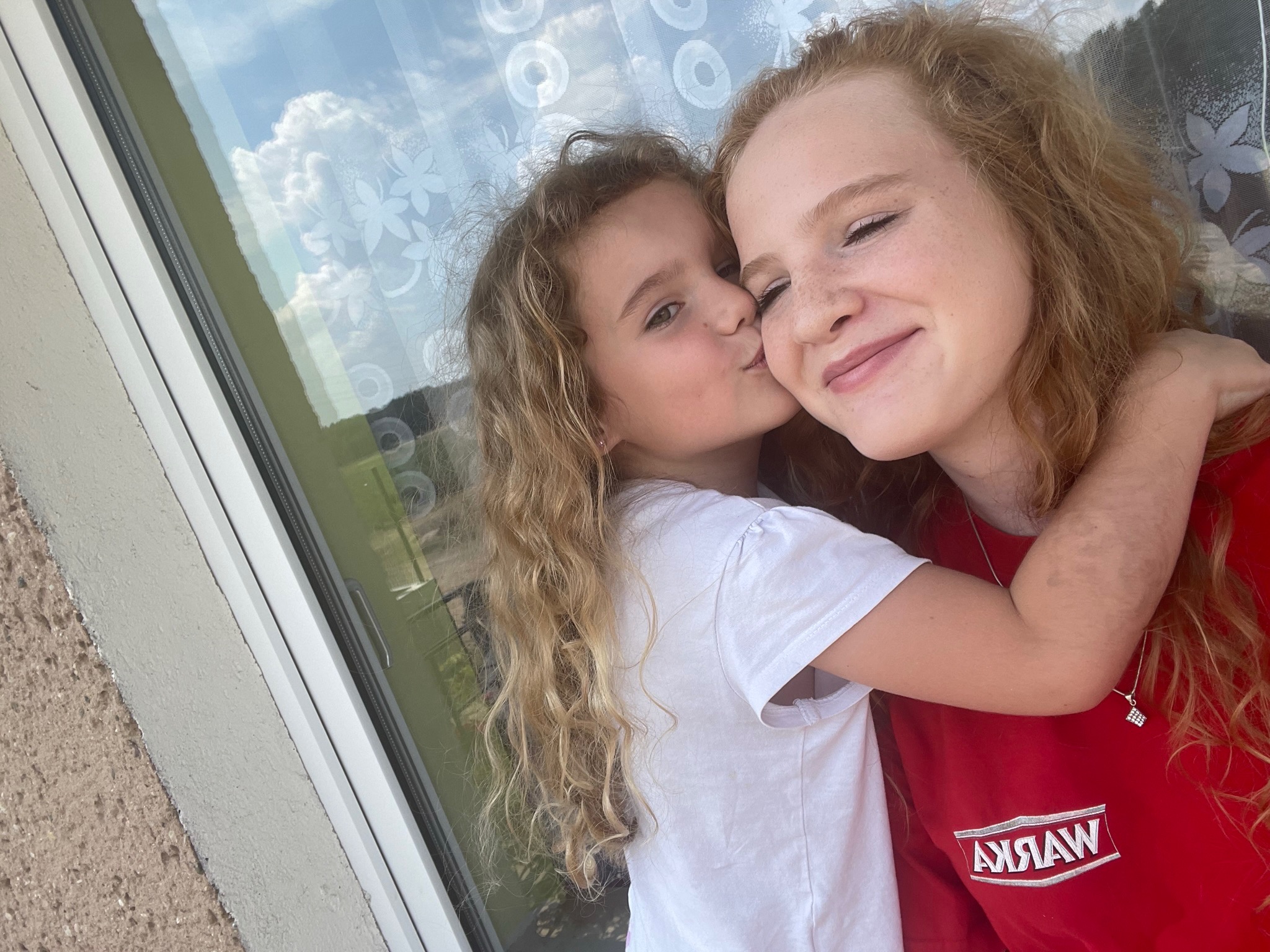 Młodsza dziewczynka całuje w policzek starszą, obie z kręconymi włosami, przytulone na tle okna z widocznymi chmurami.