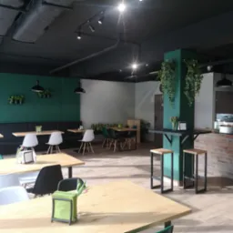 Nowoczesna aranżacja wnętrza kawiarni z drewnianymi stołami, krzesłami w stylu skandynawskim, zieloną ścianą z wiszącymi roślinami i ceglanym barem.