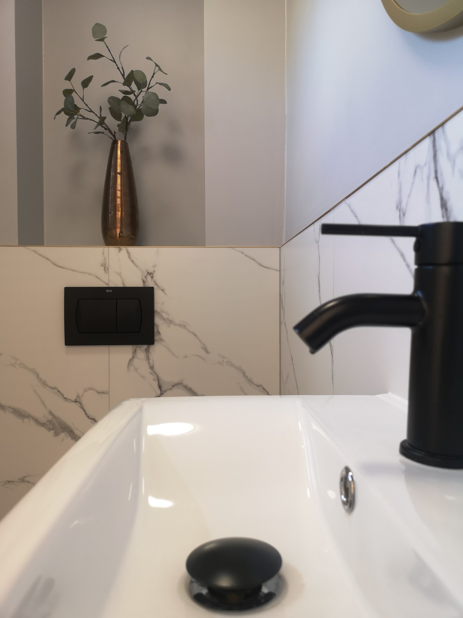 Biała umywalka z czarną armaturą na tle marmurowych płytek, dekoracyjna waza z zielonymi gałązkami, minimalistyczny design.