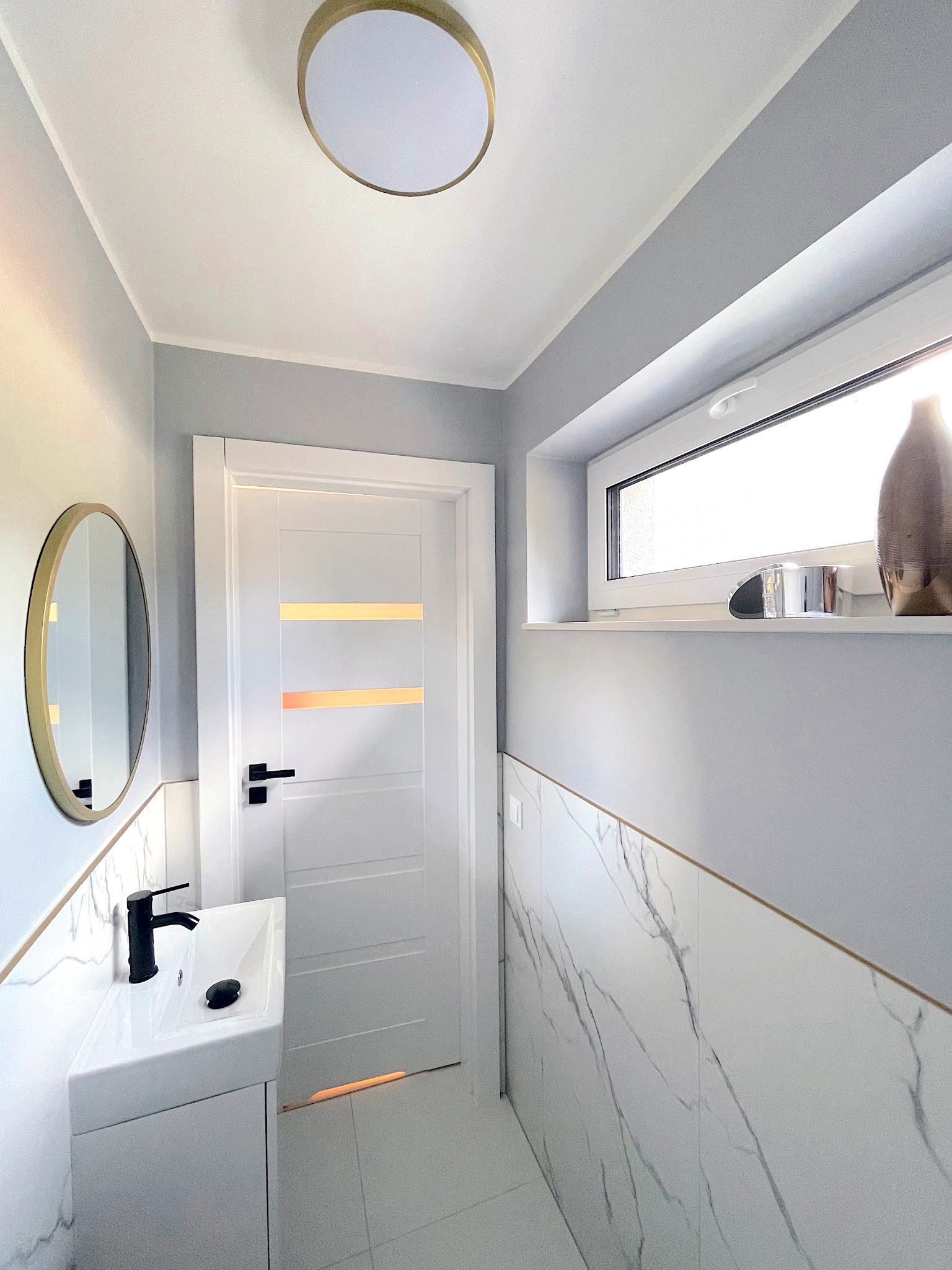 Nowoczesna łazienka w odcieniach szarości i bieli z marmurowymi płytkami, okrągłym lustrem w złotej ramie i minimalistyczną umywalką z czarną armaturą.